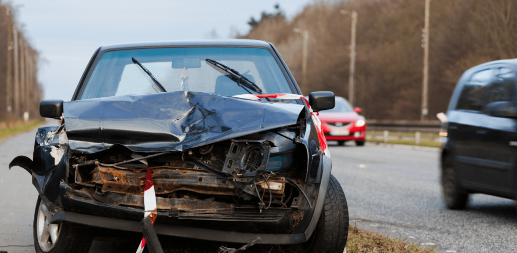 A heavily damaged car with a crushed front parked by the roadside, with other vehicles in the background