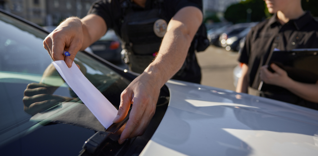 Police officer issuing a parking ticket on a car windshield while another takes notes, illustrating enforcement of parking-related traffic violations in urban areas
