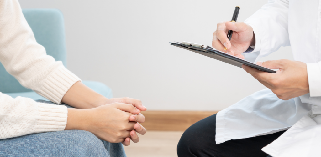 Close-up of a patient sitting with hands clasped while a doctor writes notes on a clipboard