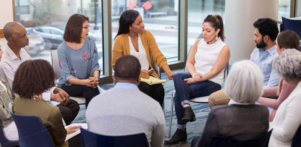 Group of people seated in a circle having a discussion in a bright room with large windows