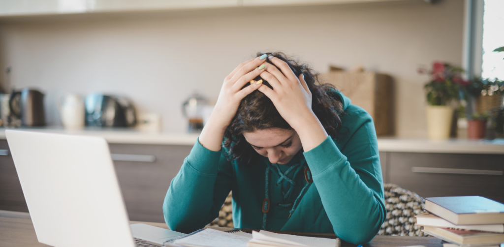 Young woman in a kitchen with her head in her hands, appearing stressed, with a laptop and notebooks on the table