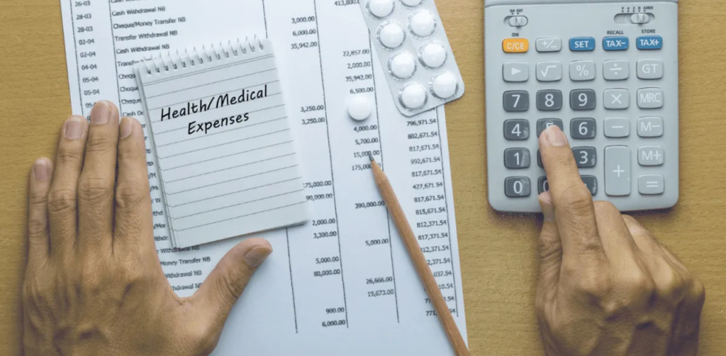 Hands calculating health or medical expenses with documents, a notepad, pills, and a calculator on a desk