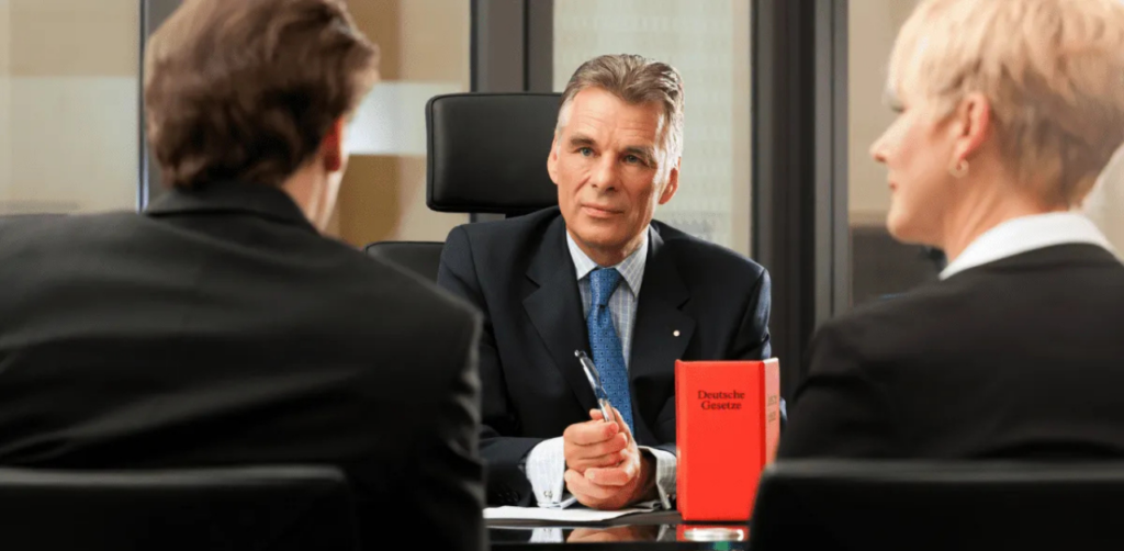 Lawyer in a suit sits at a desk holding a pen while speaking to two clients in a formal meeting. A red law book is on the table