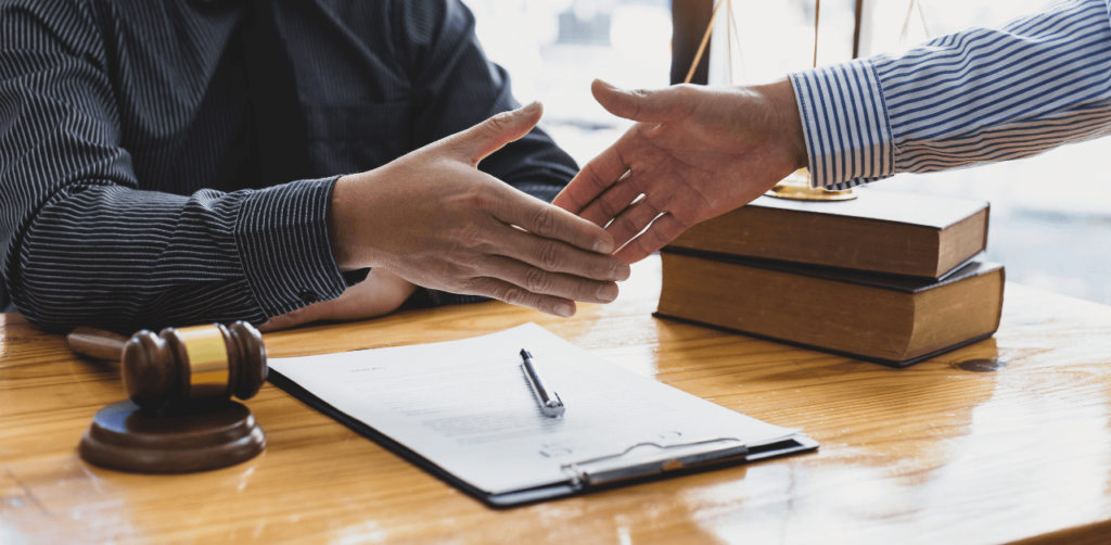 Two people shaking hands over a desk with legal documents, a gavel, and stacked books, suggesting a legal agreement