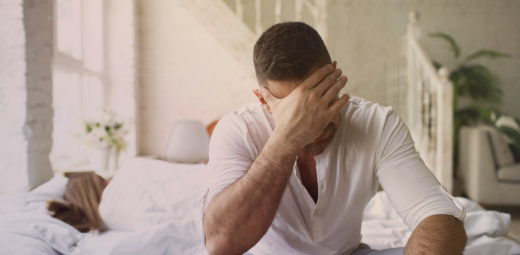 Person sits on a bed with their head in their hand, appearing distressed