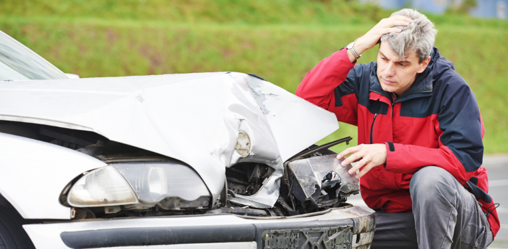 A distressed man kneeling by a car with a damaged front, inspecting the vehicle, possibly after an accident