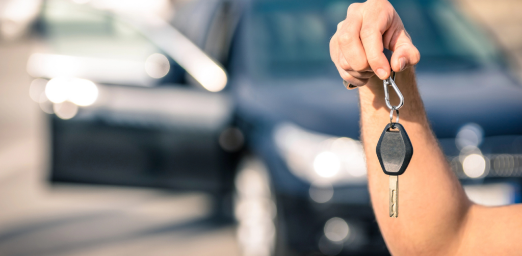 A hand holding car keys with a blurred car in the background, suggesting new ownership or car rental