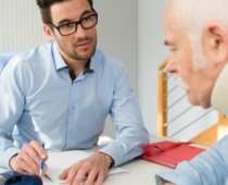 A man with glasses and a pen discusses paperwork with an elderly man wearing a neck brace, suggesting a consultation or legal advice after an injury