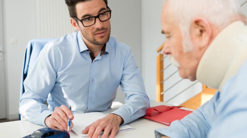 A man with glasses and a pen discusses paperwork with an elderly man wearing a neck brace, suggesting a consultation or legal advice after an injury