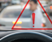 View from behind the steering wheel of a car, with a warning triangle overlay and a blurred pedestrian crossing the street ahead