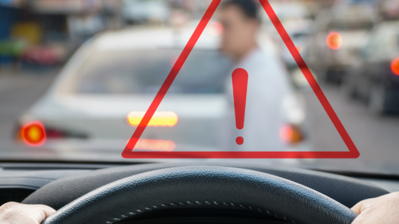 View from behind the steering wheel of a car, with a warning triangle overlay and a blurred pedestrian crossing the street ahead