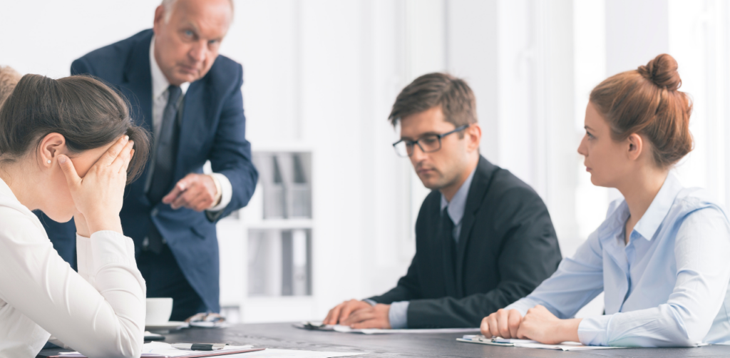 A frustrated employee with her head in her hands during a tense meeting, while an older man points at her, and two colleagues look on