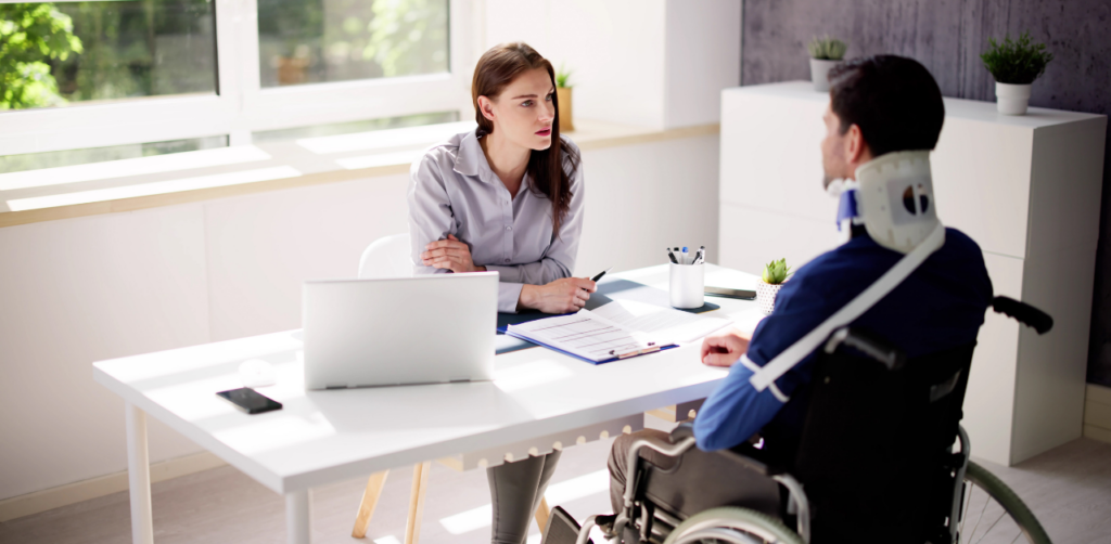 A man in a wheelchair with a neck brace sits across from a woman at a desk, possibly in a consultation or meeting setting