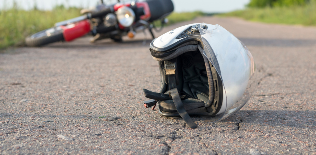 A motorcycle helmet lies on the road, with a red motorcycle in the background, both suggesting an accident scene on a rural road