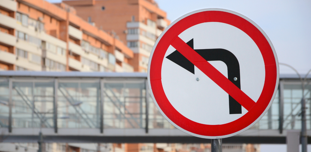 A no left turn traffic sign with apartment buildings and a pedestrian bridge in the background