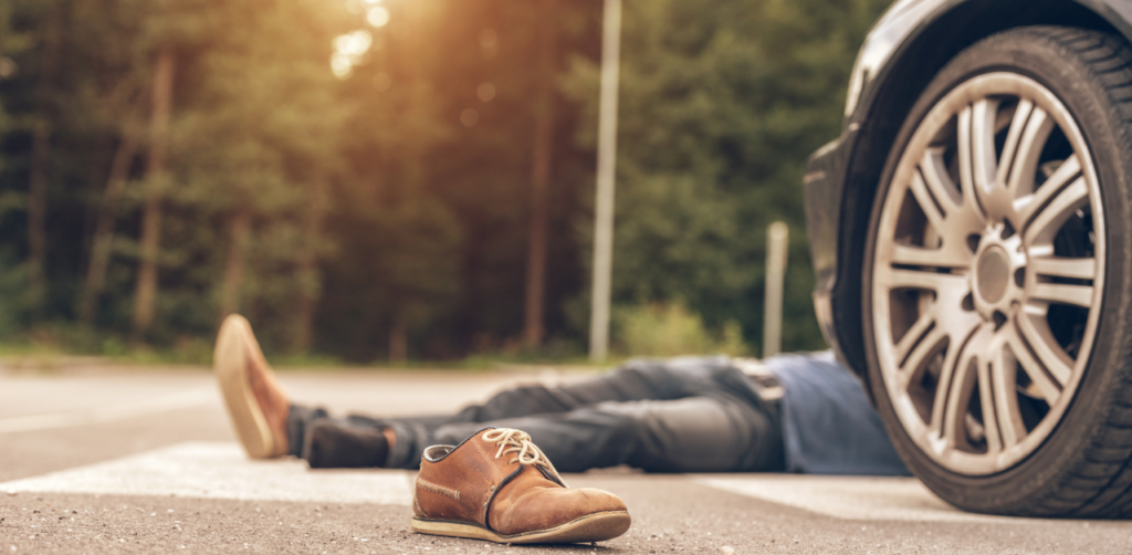 A shoe is on the road, and a person is lying on the ground near a car, indicating a pedestrian accident