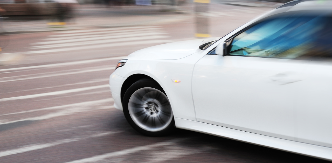 A white car driving quickly through a crosswalk, with motion blur visible on the wheels