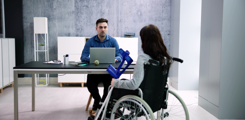 A woman in a wheelchair holds crutches while speaking to a man sitting at a desk, possibly during a consultation or interview