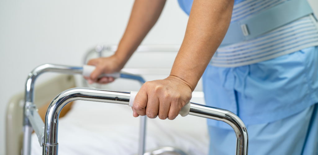 Close-up of a person using a walker, focusing on their hands gripping the frame, while standing near a hospital bed