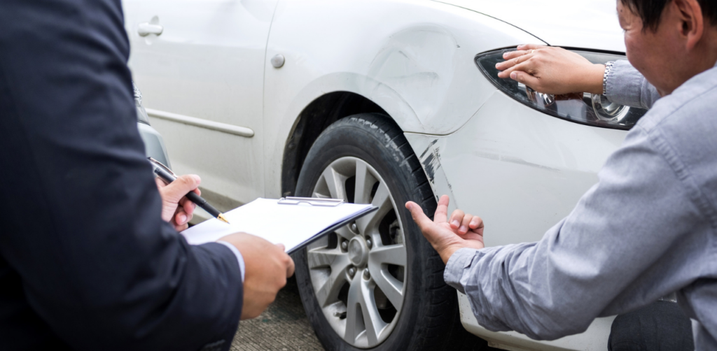 Two men examining damage to a car, one holding a clipboard while the other gestures at the scratched surface