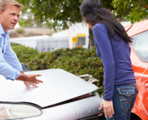 Two people stand beside their damaged cars after an accident, with one man inspecting the front of his vehicle while speaking to the other