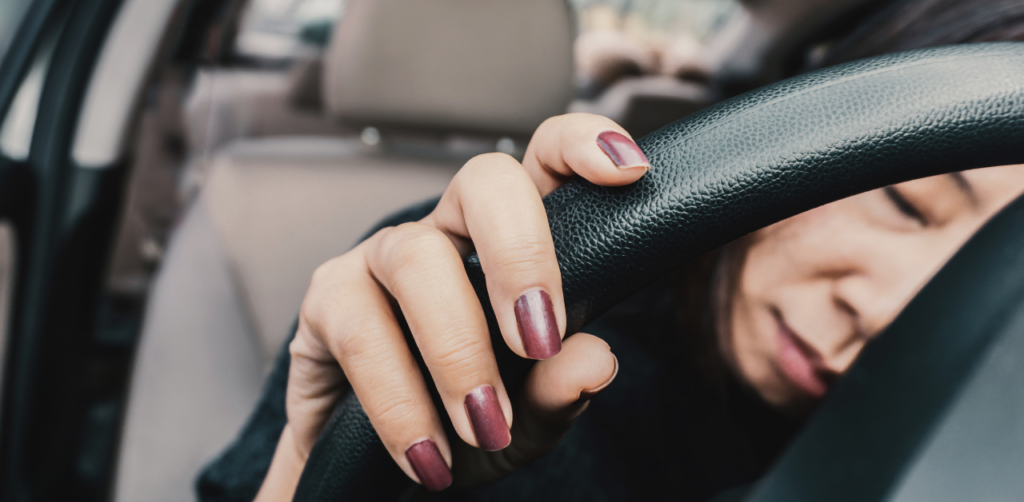 A close-up of a woman gripping a steering wheel, her eyes closed as if exhausted or overwhelmed