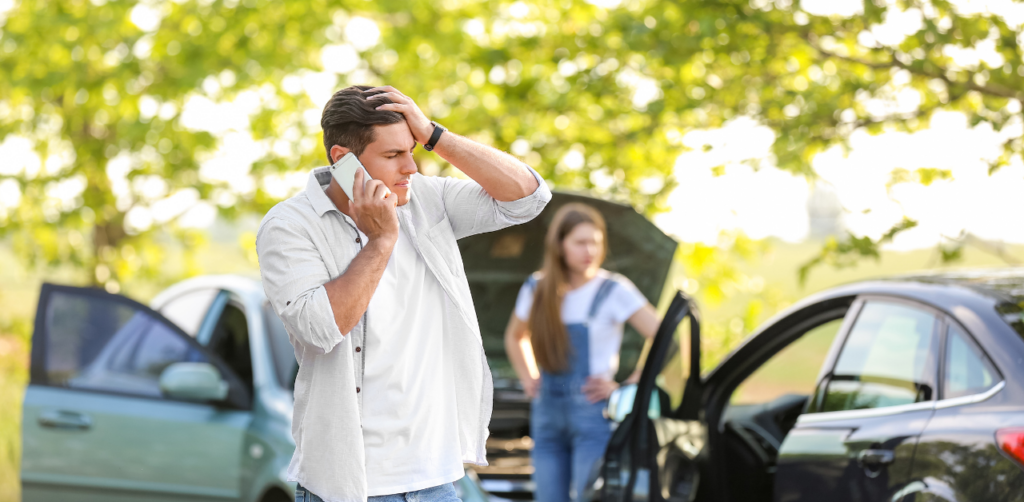 A man on the phone looking distressed after a car accident, with a woman standing near another vehicle in the background