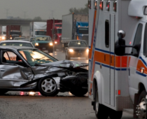 A silver sedan with severe front-end damage on a busy highway, with an ambulance and traffic in the background