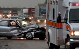 A silver sedan with severe front-end damage on a busy highway, with an ambulance and traffic in the background