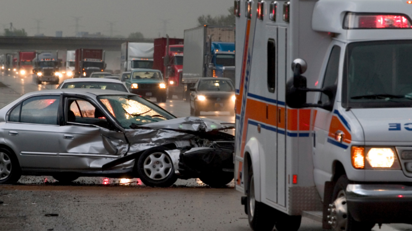 A silver sedan with severe front-end damage on a busy highway, with an ambulance and traffic in the background