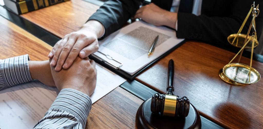 Two people in a legal office, one offering comfort with clasped hands, next to a gavel and scales of justice on a desk