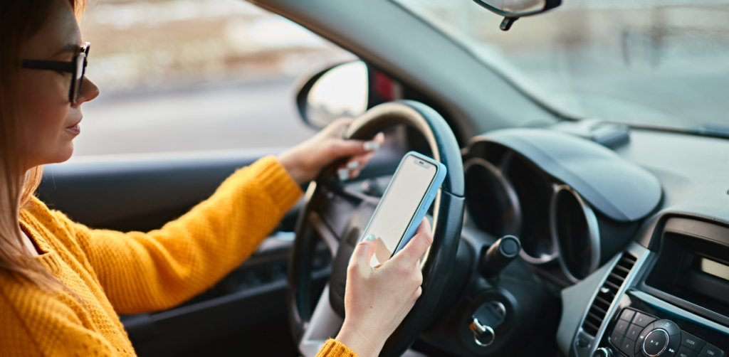 Woman in a yellow sweater inside a car, holding a smartphone while driving, with her other hand on the steering wheel