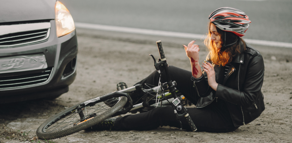 Cyclist wearing a helmet sits injured next to a fallen bike, examining a bloody arm near a car on the roadside