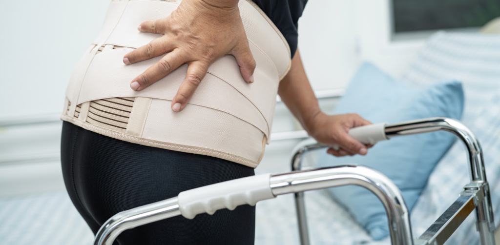 Injured person in back brace uses walker, hand on lower back showing pain from injury