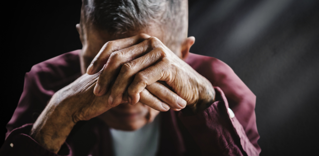 Elderly man clasps hands over face in emotional distress, sitting alone in a dark, moody setting