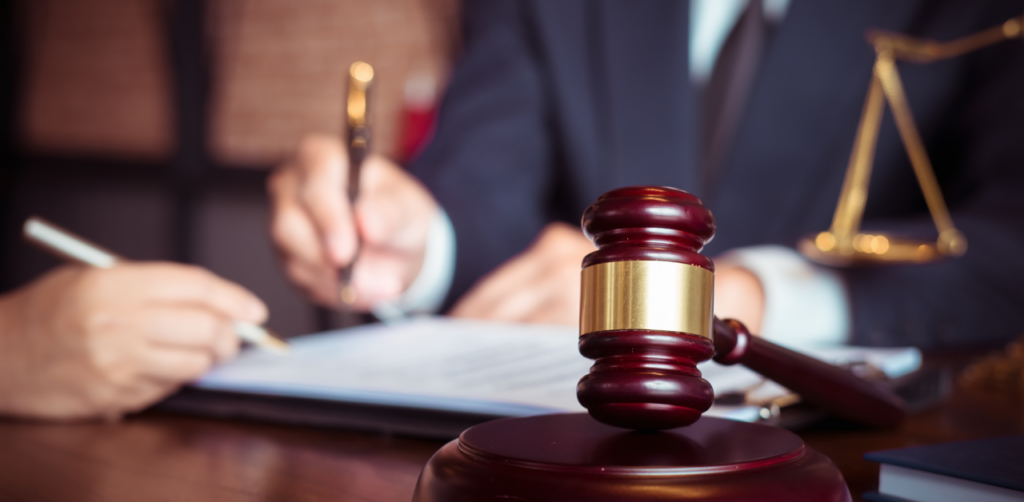 Close-up of a judge's gavel on a desk with blurred figures in suits reviewing legal documents in the background