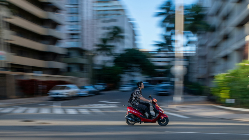 Person riding a red scooter through a city street with motion blur, representing fast urban commuting and mobility