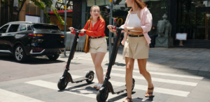 Two women walking electric scooters across a city crosswalk, highlighting urban micromobility and shared transportation