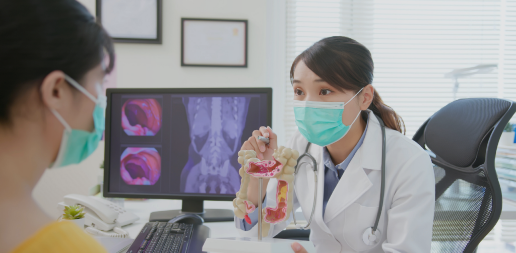 Doctor wearing a face mask explaining a digestive system model to a patient in a medical office with scan images displayed on a monitor