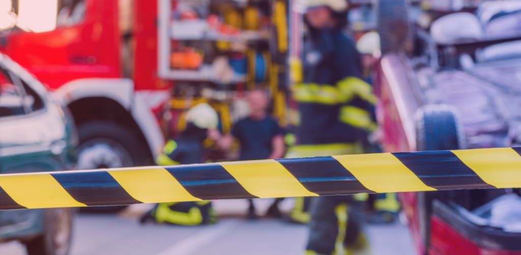 Emergency response scene with caution tape in the foreground and firefighters working near damaged vehicles in the background