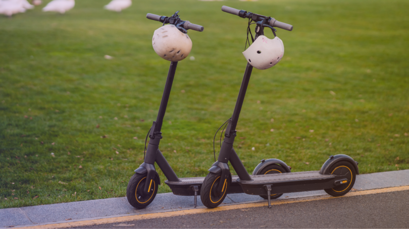 Two electric scooters with helmets hanging from the handlebars parked beside a grassy area