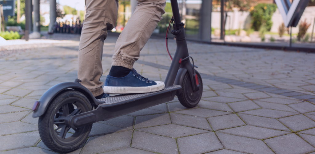 Close-up of a person standing on an electric scooter, with one foot on the deck and the scooter positioned on a paved urban walkway