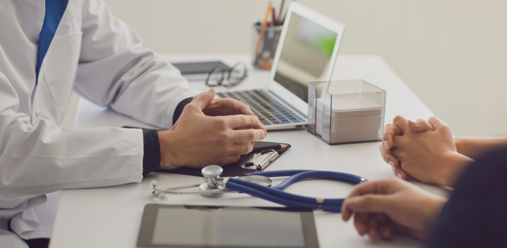 Doctor consulting with two people across a desk in a medical office, with a stethoscope, clipboard, and laptop visible during the discussion