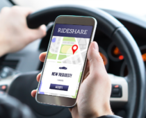 Driver holding a smartphone displaying a rideshare app with a new ride request while seated behind the wheel of a car
