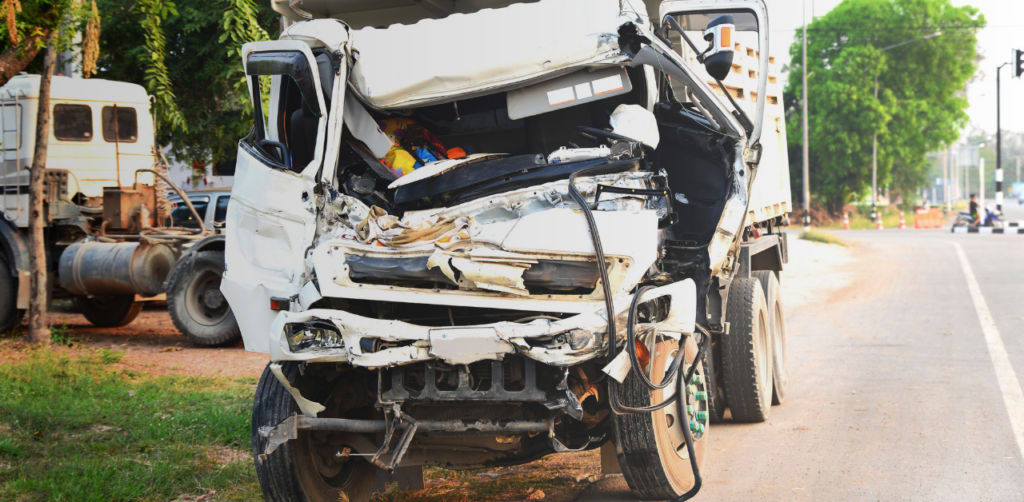 Front view of a severely damaged box truck parked beside a road, showing extensive crash damage to the cab and front end after a collision