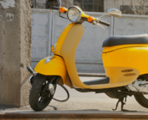 Yellow scooter parked on a roadside curb beside a concrete utility pole, with a street and parked car in the background