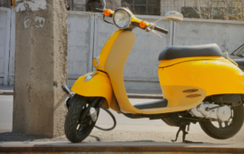 Yellow scooter parked on a roadside curb beside a concrete utility pole, with a street and parked car in the background