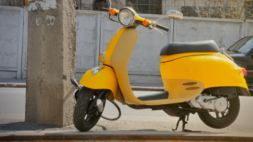 Yellow scooter parked on a roadside curb beside a concrete utility pole, with a street and parked car in the background