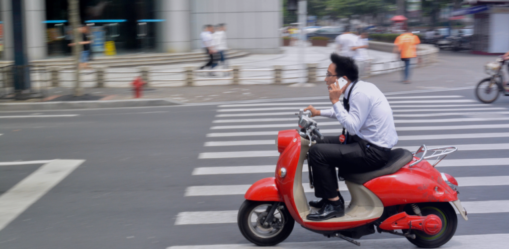 Person riding a red scooter through a city intersection while holding a cellphone to their ear, creating a distracted-driving safety hazard
