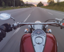 First-person view from a motorcycle traveling on a multi-lane highway at dusk, with the rider’s gloved hands on the handlebars and open road ahead